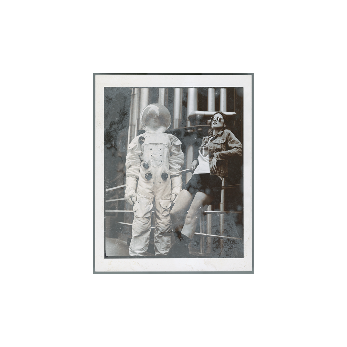 Black and white photo print of a person in a full astronaut suit standing beside another individual in casual clothing, who is seated on a metal railing. Behind them, an array of large industrial pipes.